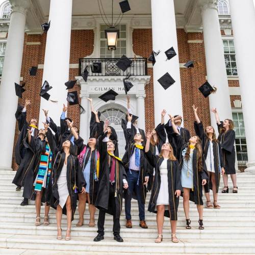 A group of 15 students in graduation regalia throw their caps in the air while standing on the white marble steps in front of Gilman Hall