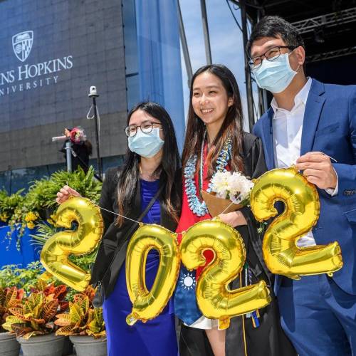 Three students, two females and one male, hold gold balloons in the shape of the number 2022