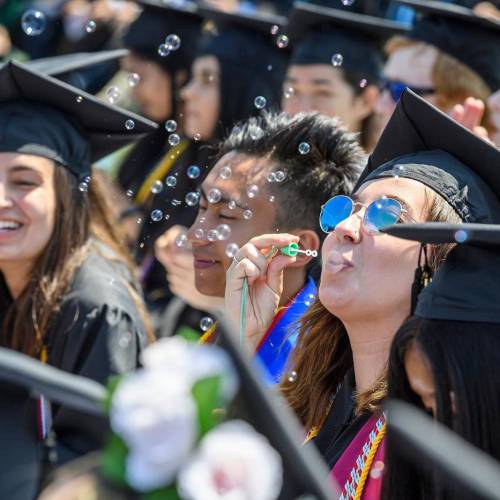 A student wearing a graduation cap and sunglasses blows dozens of small bubbles into the air while nearby students nearby smile