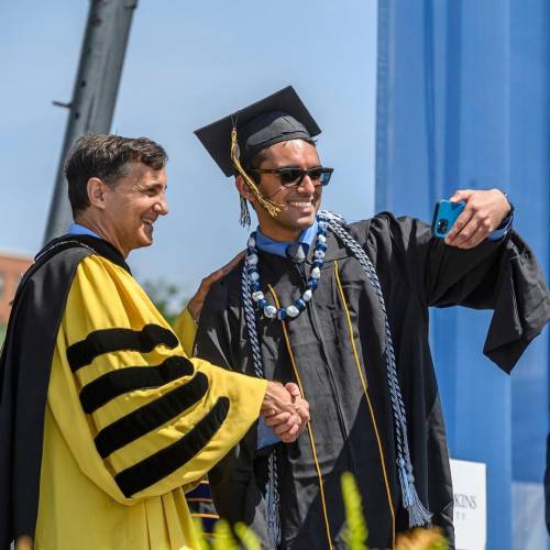 A graduating student wearing sunglasses takes a selfie while shaking hands with JHU President Ron Daniels
