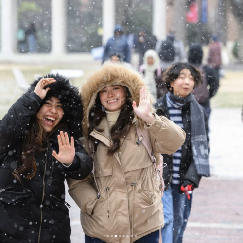 Two girls with winter coats and hoods smile while light snow falls around them.